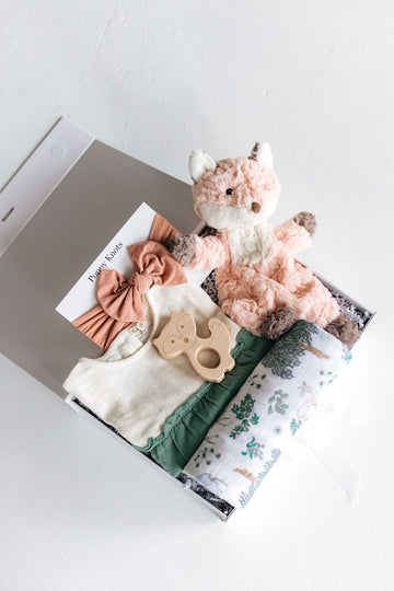 baby girl gift set including a white and green dress, forest pattern swaddle, fox shaped wood teether, fox lovey and blush headband