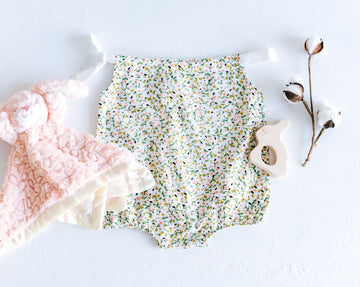 flatlay photo of pink bunny blanket, floral bubble romper with yellow and green flower buds, and bunny shaped wood toy