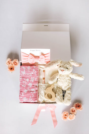 baby girl gift box with pink floral swaddle, pink bow headband, white dress, bunny shaped wood teether, and bunny plushie